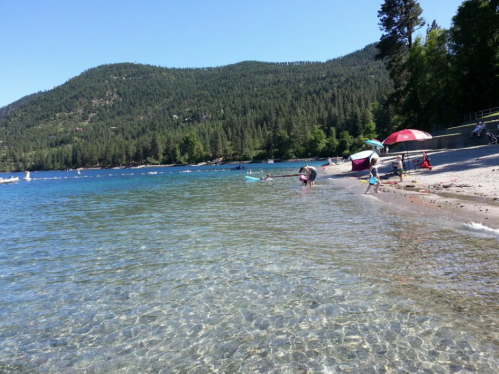 A sunny beach scene with clear water, people enjoying the shore, and green mountains in the background.