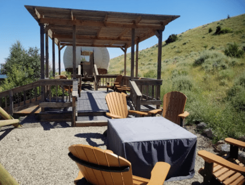 A rustic outdoor seating area with wooden chairs and a table, overlooking a grassy hillside and a wagon structure.