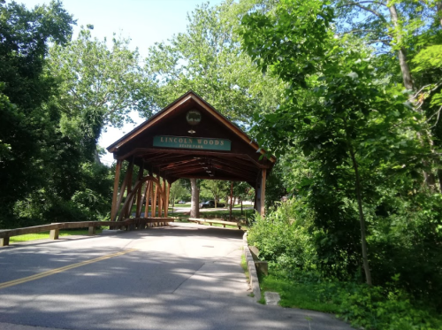A wooden covered bridge with "Lincoln Woods" sign, surrounded by lush green trees and a paved road.