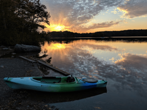 A kayak rests on a rocky shore at sunset, reflecting vibrant colors on the calm water.