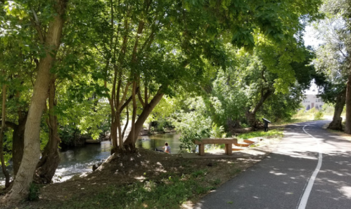 A peaceful path alongside a river, shaded by trees, with a bench and a person fishing in the water.