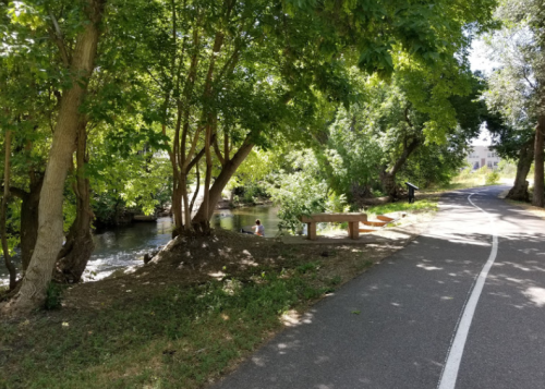 A peaceful path alongside a river, shaded by trees, with a bench and a person sitting by the water's edge.