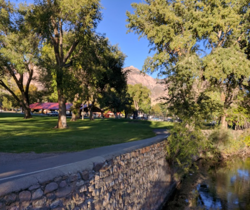 A scenic park with trees, a stone wall, and a river, set against a backdrop of mountains under a clear blue sky.