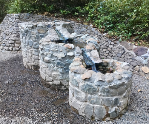 Three stone water fountains with flowing water, surrounded by gravel and greenery.