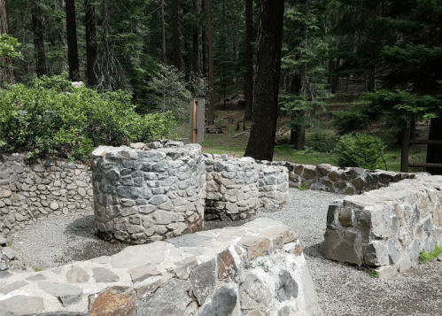 Stone well structure surrounded by trees in a forested area, with gravel paths and greenery nearby.