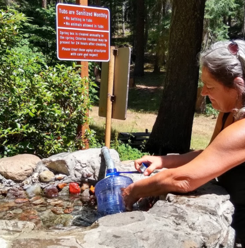 A woman fills a water container at a natural spring, with a sign about sanitation and safety in the background.