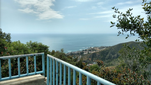 A scenic view of the ocean and coastline from a hillside, framed by greenery and a blue railing in the foreground.