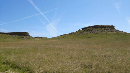A wide grassy landscape with two rocky outcrops under a clear blue sky with wispy clouds.