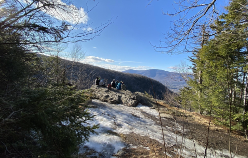 A group of hikers sits on a rocky outcrop, surrounded by trees, overlooking a scenic mountain view on a clear day.