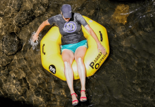 A person in a yellow inner tube splashes in clear water, wearing a cap and casual summer attire.