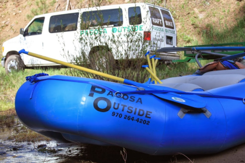 A blue raft is parked by a river, with a van in the background advertising rafting and biking services.