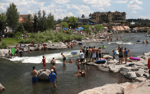 A lively river scene with people swimming, floating on tubes, and relaxing on rocks, surrounded by greenery and buildings.