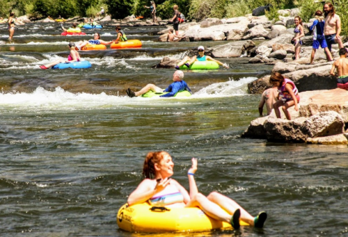 People enjoying a sunny day tubing on a river, with some relaxing on inflatable rafts and others playing by the shore.