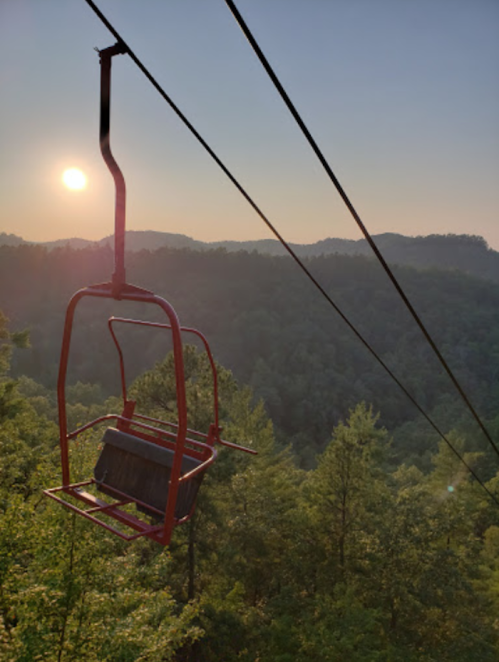 A red chairlift hangs over a lush green landscape at sunset, with mountains in the background.