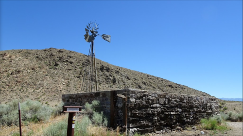 A weathered windmill stands atop a stone base, surrounded by arid hills under a clear blue sky.