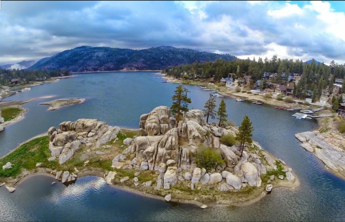 Aerial view of a rocky island surrounded by a lake, with trees and mountains in the background under a cloudy sky.