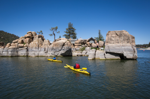 Two kayakers in yellow kayaks paddle near rocky formations on a calm lake under a clear blue sky.