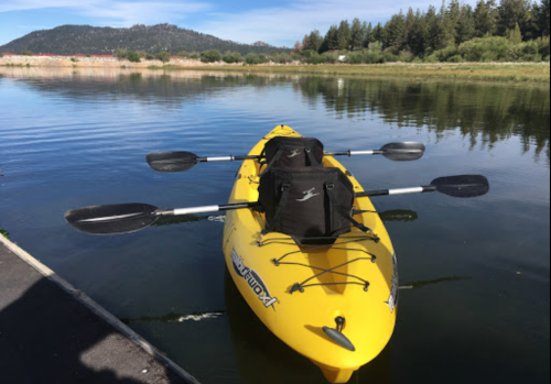 A yellow kayak with two paddles rests on calm water, surrounded by trees and mountains in the background.