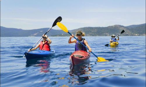 Three people kayaking on a calm lake, surrounded by mountains and clear blue skies.
