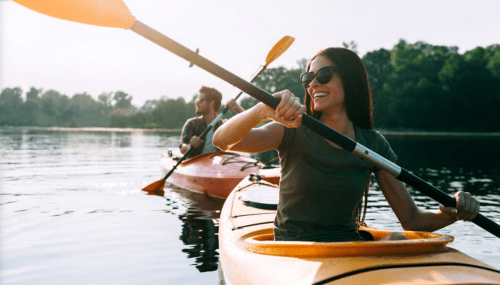 A woman in a kayak smiles while paddling on a calm lake, with another kayaker in the background.