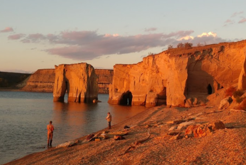 Two people stand on a sandy shore near towering rock formations at sunset, reflecting on the calm water.