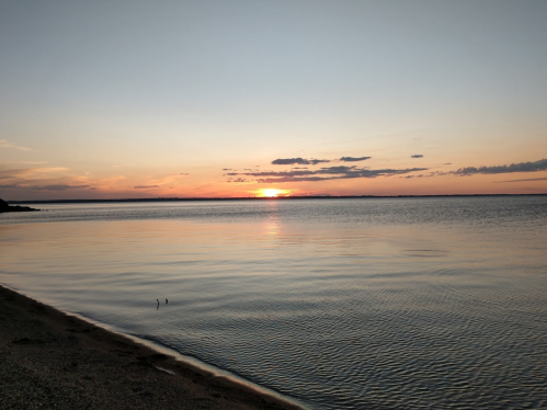 A serene sunset over calm waters, with soft clouds and a sandy shore in the foreground.