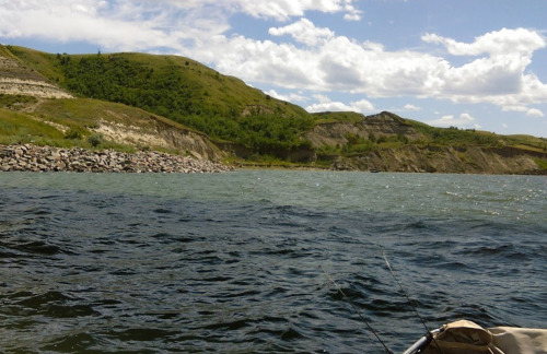 A serene view of a river with rolling green hills and a rocky shoreline under a partly cloudy sky.