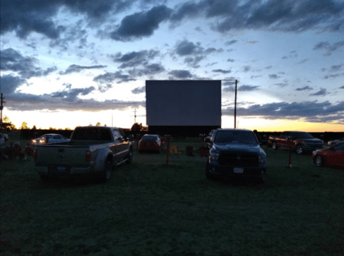 A drive-in movie theater at dusk, with parked cars facing a large blank screen against a colorful sky.