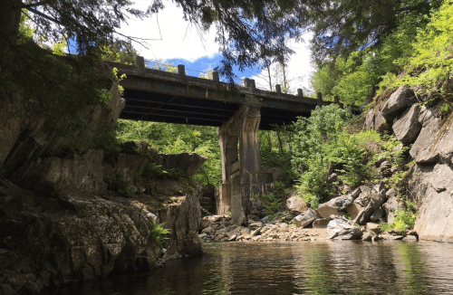 A bridge spans a rocky river, surrounded by lush greenery and blue skies with scattered clouds.