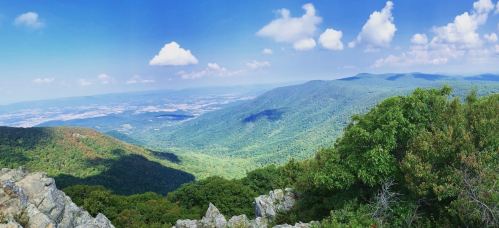 A panoramic view of lush green mountains under a bright blue sky with fluffy clouds.