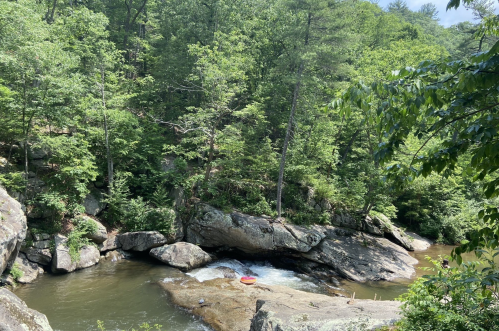A serene river flows through a lush green forest, with large rocks and a small kayak visible in the water.