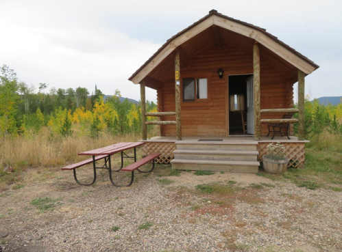 A rustic wooden cabin with a porch and picnic table, surrounded by trees and open grassland.