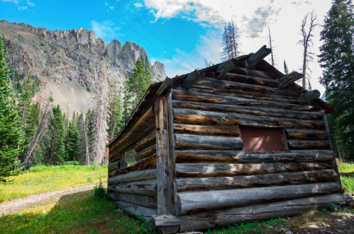A rustic log cabin surrounded by tall trees and mountains under a bright blue sky.