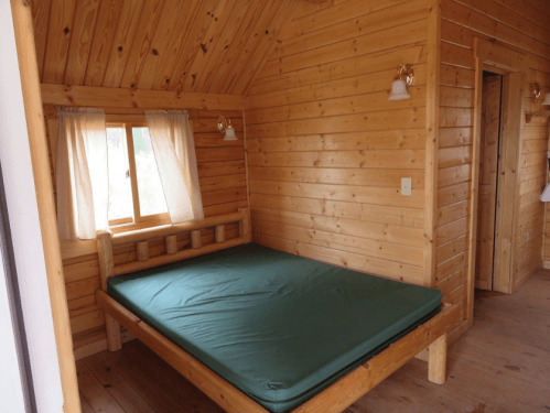 Cozy wooden cabin interior featuring a simple bed, natural wood walls, and a window with sheer curtains.