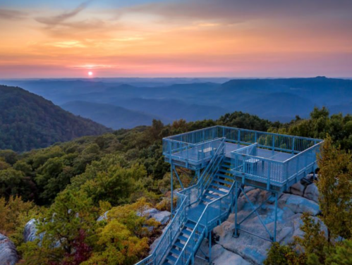 A metal observation deck overlooks rolling mountains at sunset, with vibrant colors in the sky and lush greenery below.