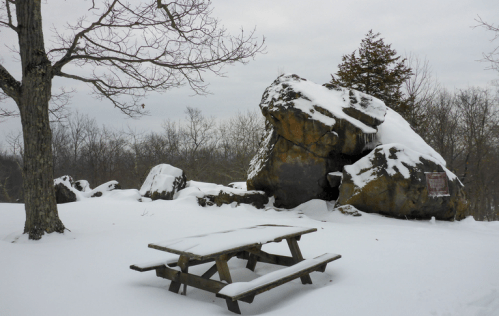 A snowy landscape featuring a picnic table and large boulders, with bare trees in the background under a cloudy sky.