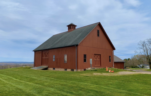 A red barn with a sloped roof sits on green grass, surrounded by rolling hills under a cloudy sky.
