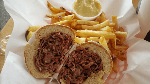 A sandwich filled with sliced meat, served with fries and a small cup of mustard on a paper-lined plate.