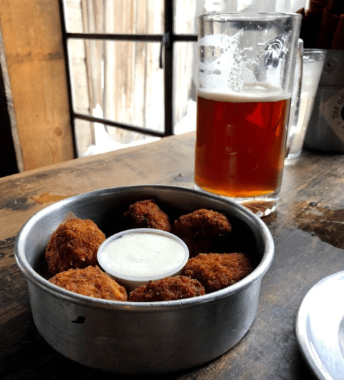 A metal bowl of golden-brown falafel balls with a small cup of dipping sauce, next to a glass of amber beer.