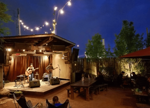 Two musicians perform on a wooden stage under string lights, with an outdoor audience seated nearby in a cozy setting.