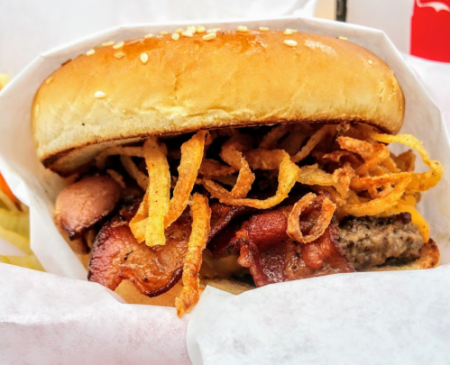 A close-up of a burger with bacon, crispy onions, and a sesame seed bun, served in a paper wrapper.