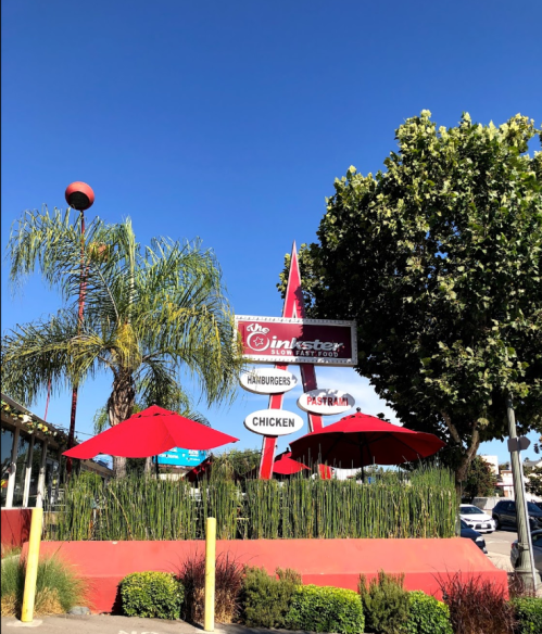 A retro diner sign with palm trees and red umbrellas against a clear blue sky.
