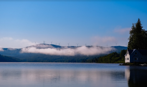 A serene lake reflects a clear blue sky, with mountains and wind turbines in the background, partially shrouded in mist.