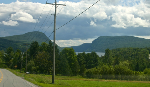 Scenic view of mountains and a lake under a cloudy sky, with a road and power lines in the foreground.