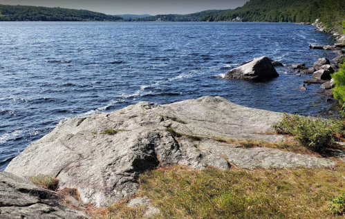 A rocky shoreline by a lake, with gentle waves and green hills in the background under a clear sky.