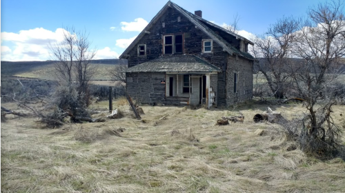 Abandoned two-story house with a weathered exterior, surrounded by dry grass and sparse trees under a blue sky.