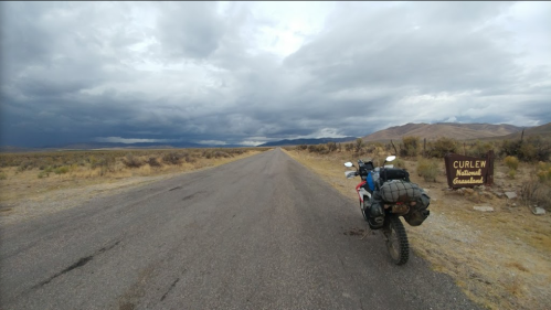 A motorcycle parked beside a road leading to Curlew National Grassland, with dark clouds overhead and mountains in the distance.