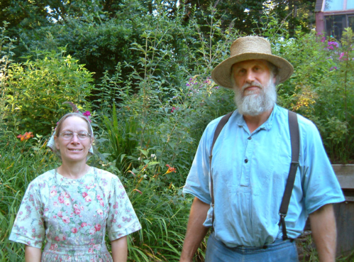 A man and woman stand together in a lush garden, surrounded by colorful flowers and greenery.