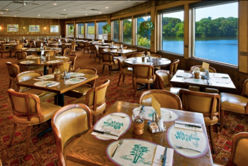 Cozy restaurant interior with wooden tables, chairs, and a view of a lake through large windows.