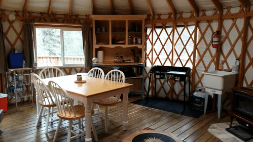 Cozy yurt interior with a wooden dining table, chairs, kitchenette, and large window letting in natural light.